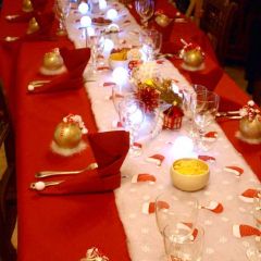 Table harmonie de rouge, blanc et touches de doré composée d'une nappe rouge, d'un chemin de table imitation neige avec lumières, de serviettes rouges bottes de lutin, de boîtes boules de Noël et de centres de table végétaux et figurines.